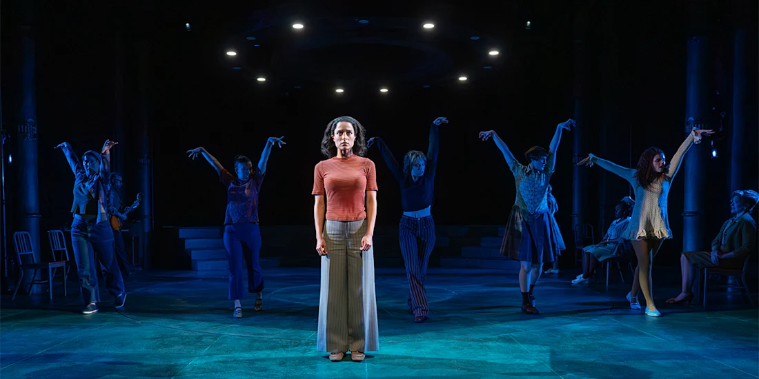 A woman stands center stage under a spotlight while six other actors pose dramatically behind her, all set against a dark, blue-lit theatrical background.