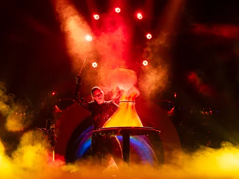 A performer in a sparkling outfit stands behind a table with a large, conical flask emitting colored smoke, under dramatic red stage lighting.