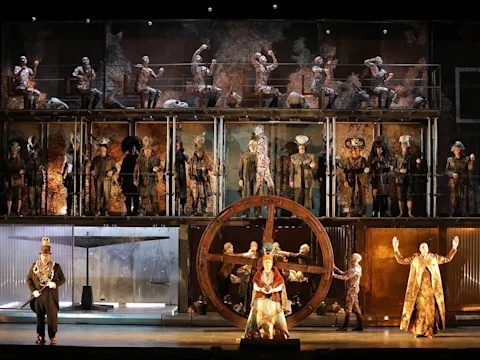 Production photo of Akhnaten in Los Angeles, showing a stage performance with multiple actors in elaborate costumes, set across two levels; a large wooden wheel is positioned center stage with dramatic lighting.
