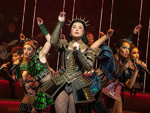 Production photo of SIX in Costa Mesa showing performers in colorful, ornate costumes sing and dance on stage under dramatic lighting, with one person standing at the center holding a microphone.