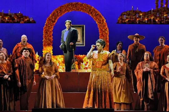 A group of opera performers in colorful costumes sings on stage with marigold decorations, candles, and an arch in the background.