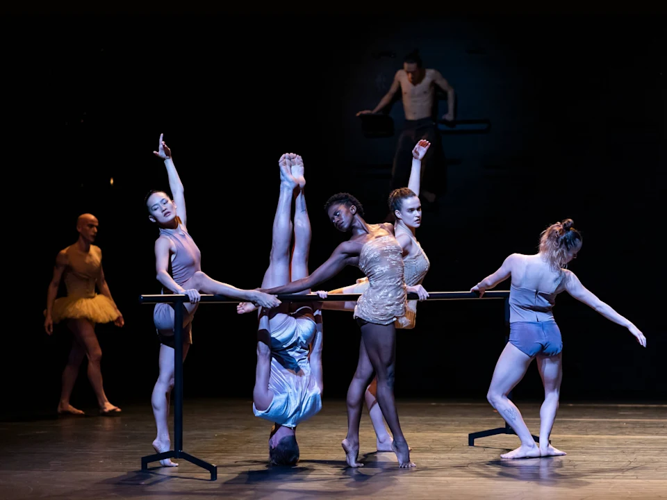 Dancers perform on stage around a ballet barre, with one dancer upside down. Others stand in varied positions, wearing different costumes. A person in a tutu observes from the background.