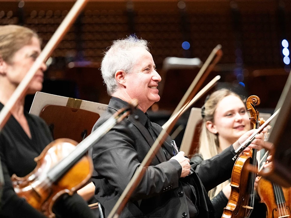 Several musicians in formal black attire play string instruments in an orchestra, with music stands in front of them. One musician in the center is smiling.