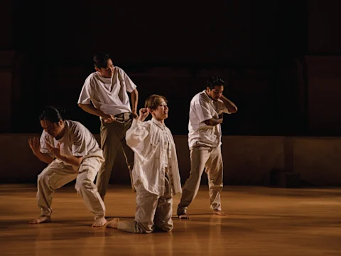 Four people wearing loose, white clothing perform expressive movements on a wooden floor, with one person kneeling and the others standing in a dimly lit space.