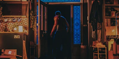 A person in a tank top stands in an open doorway at night, illuminated by blue light from outside. The room is warmly lit and cluttered with household items and hanging cups.