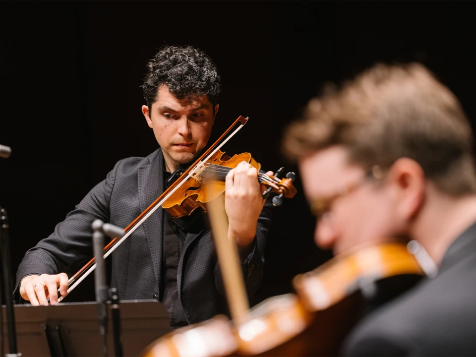 Two musicians playing string instruments, focused on their performance. One is in the foreground, slightly out of focus, and the other is in the background, holding a violin.