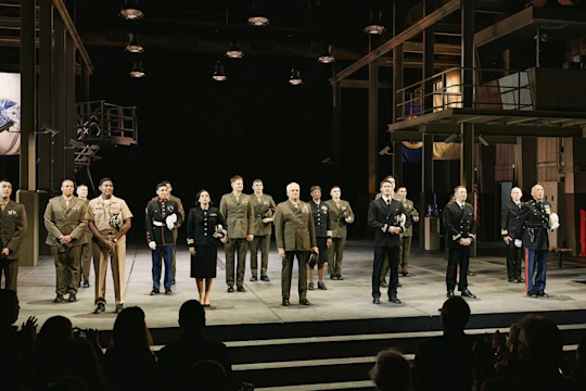 A group of military personnel in formal uniforms standing in a line on a theater stage, facing an audience.