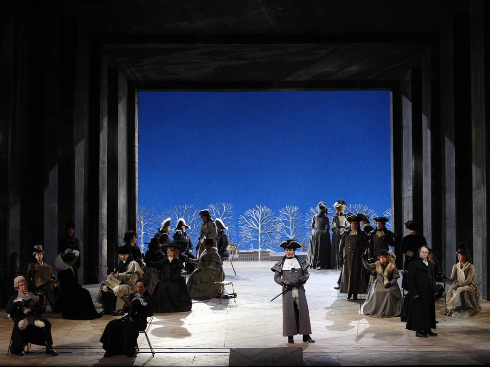 A stage performance with actors in period costumes. A central figure stands in the foreground, while others are seated or standing in the background against a blue backdrop with tree silhouettes.