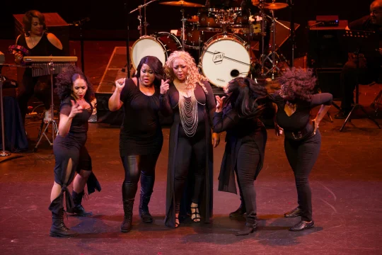 A group of five women in black outfits perform a dance routine on stage with musical instruments and performers in the background.