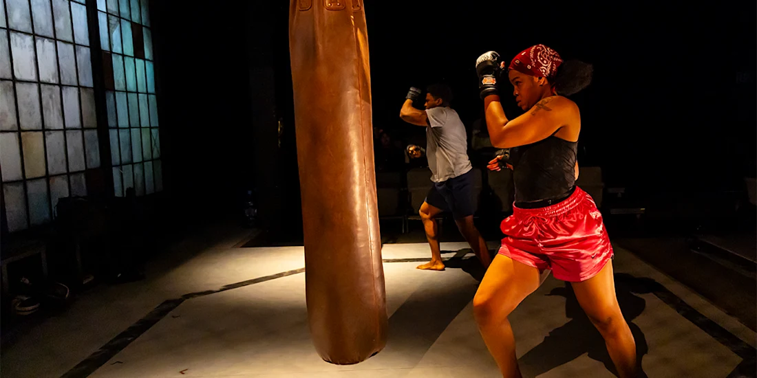 Two people practice boxing in a dimly lit gym, both wearing gloves and facing a large brown punching bag.