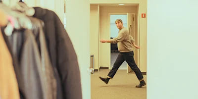 A person in a sweater and boots walks briskly through an office hallway, with coats hanging on the left and open doors in the background.