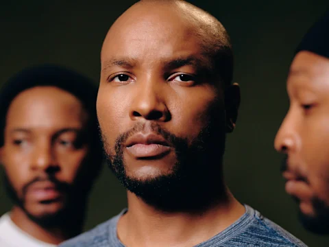 Three men stand close together, with the man in the center looking directly at the camera while the other two look away. The background is dark and out of focus.