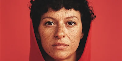 A person with short curly hair, freckles, and a red hood stares directly at the camera against a solid red background.