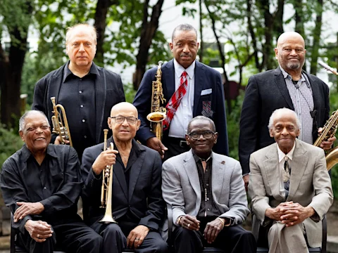 Seven older men, some holding brass and woodwind instruments, pose outdoors in suits and dress shirts, with trees in the background.