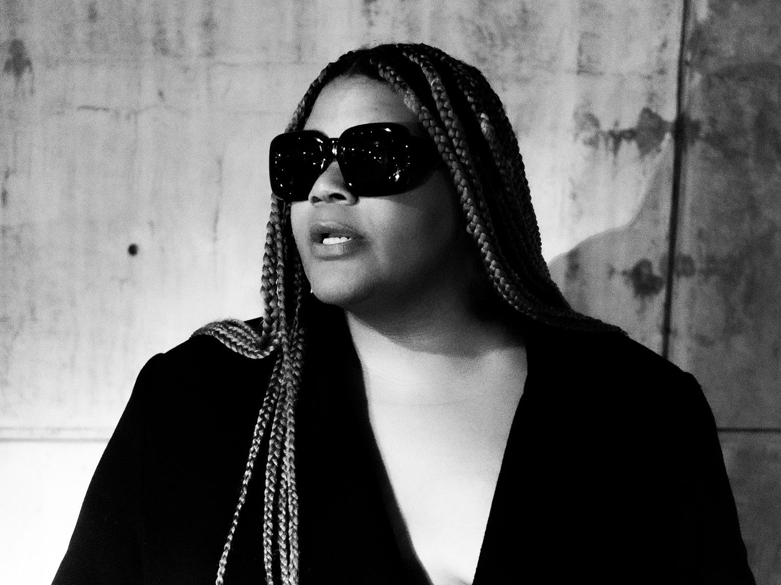 Person with long braids wearing dark sunglasses and a black outfit, standing in front of a textured wall, lit dramatically in black and white.