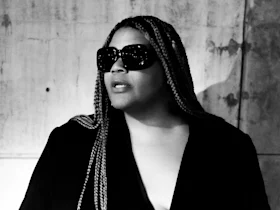 Person with long braids wearing dark sunglasses and a black outfit, standing in front of a textured wall, lit dramatically in black and white.