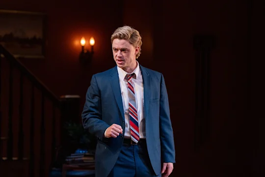 A person in a blue suit and striped tie stands indoors, gesturing with one hand, with a staircase and lit wall sconces in the background.