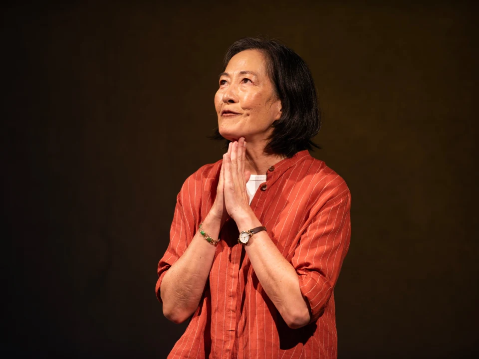 An older woman with short black hair, wearing an orange blouse, stands with hands clasped together near her face against a dark background.