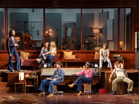 Seven people in 1970s-style clothing sit and stand around a music studio set with recording equipment, instruments, and wood-paneled walls.