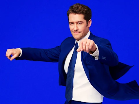 A man in a blue suit and tie poses with arms extended against a solid blue background.