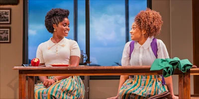 Two young women in patterned skirts sit at a table with food, smiling at each other in a brightly lit room with large windows and framed photos on the wall.