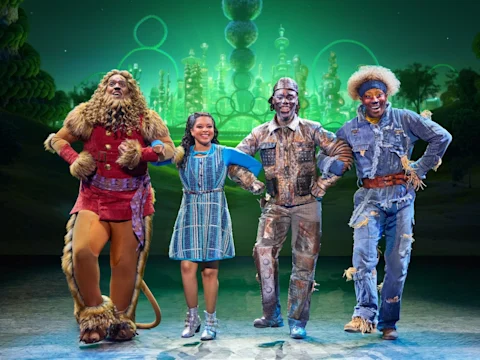 Four actors in colorful, whimsical costumes stand arm in arm on stage, with a glowing green city backdrop, portraying characters from "The Wiz.