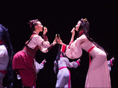 Two dancers in traditional attire face each other, holding small round mirrors and touching their faces, while performers in similar costumes stand in the background on a dark stage.