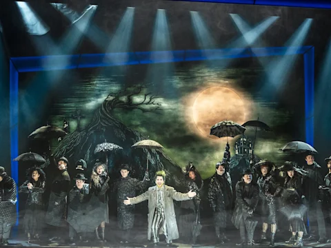 A stage performance with actors in dark costumes holding umbrellas, standing before a painted backdrop of a spooky tree and a large full moon.