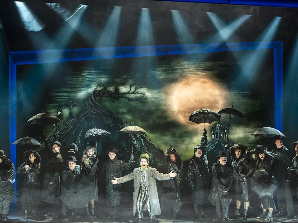 A stage performance with actors in dark costumes holding umbrellas, standing before a painted backdrop of a spooky tree and a large full moon.