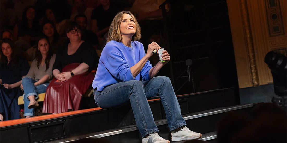 A woman in a blue sweatshirt and jeans sits on stage steps, smiling and holding an object, with an audience seated behind her.