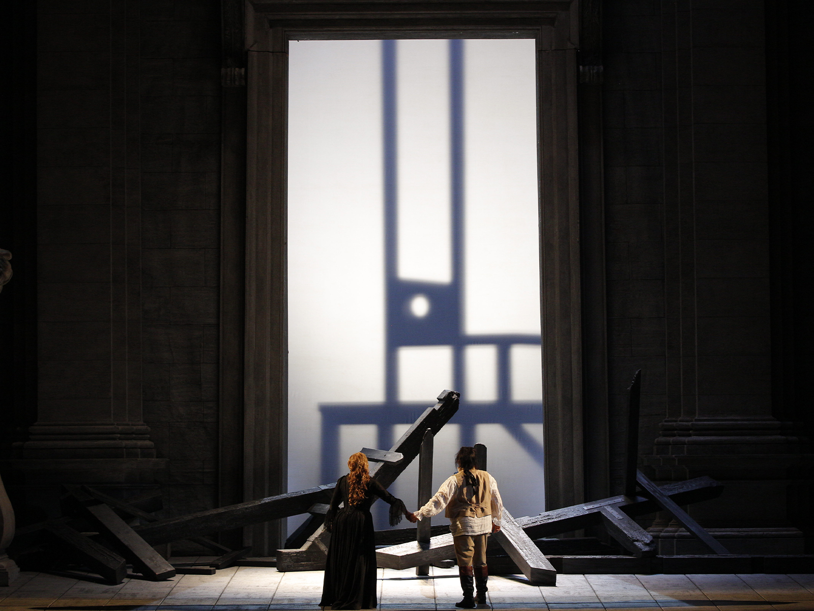 Two people stand hand in hand facing a large, illuminated doorway, surrounded by scattered wooden beams and dramatic shadows on the wall.