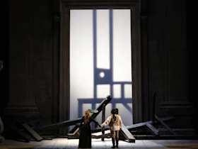 Two people stand hand in hand facing a large, illuminated doorway, surrounded by scattered wooden beams and dramatic shadows on the wall.