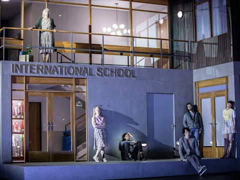 Production photo of Innocence in New York, showing seven people are positioned outside and on the balcony of a building labeled "International School," each standing or sitting apart from one another.
