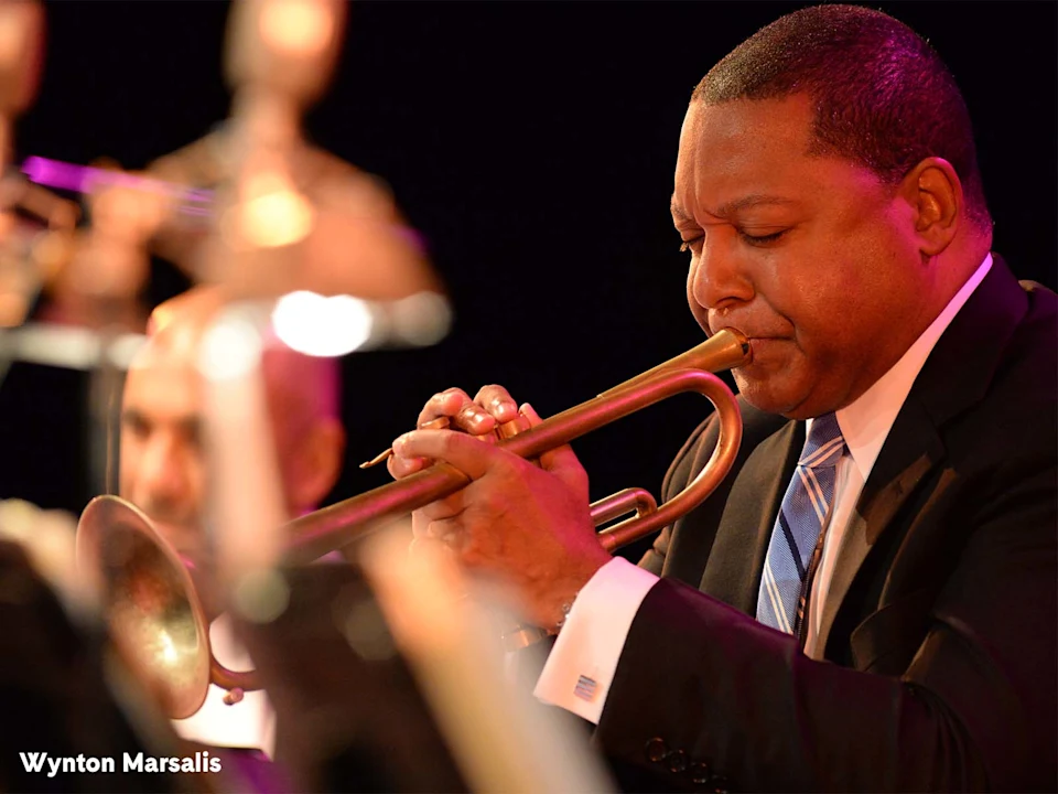 A man in a suit plays a trumpet on stage; other musicians and instruments are blurred in the foreground. Text reads "Wynton Marsalis" in the bottom left corner.