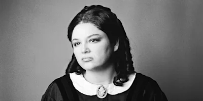 A woman with long, dark, wavy hair wearing a dark dress with a white collar and a cameo brooch, looking slightly off camera against a plain background.