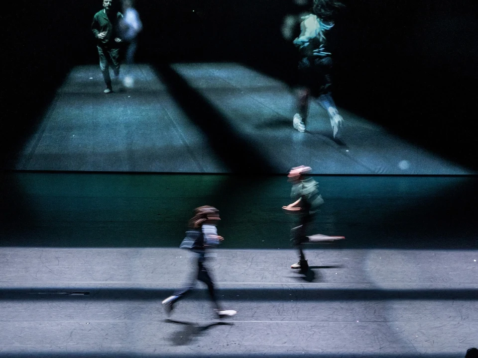 Two people run on a dark stage with blurred reflections on a screen behind them, creating a dynamic motion effect.