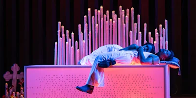 Two people dressed in white lie close together on a pink-lit altar surrounded by tall lit candles, with text inscribed on the front of the altar.