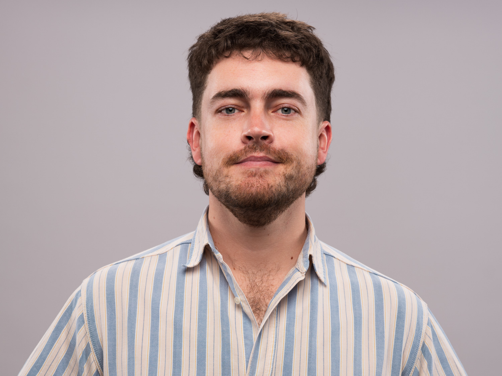 A man with short brown hair and a beard is wearing a striped button-up shirt and looking at the camera against a plain background.