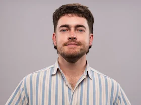 A man with short brown hair and a beard is wearing a striped button-up shirt and looking at the camera against a plain background.