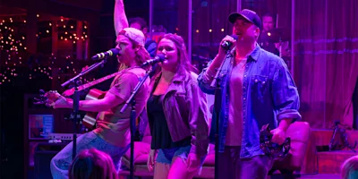 Three musicians perform on stage under purple lighting; one plays guitar, one sings, and one holds a tambourine. A drum set and colorful string lights are visible in the background.