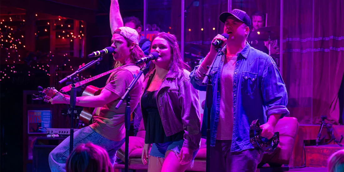 Three musicians perform on stage under purple lighting; one plays guitar, one sings, and one holds a tambourine. A drum set and colorful string lights are visible in the background.