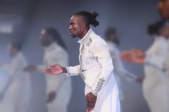 A man dressed in an embroidered white outfit stands in profile with his arm extended, while others in similar attire are blurred in the background.