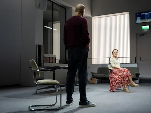 A woman in a floral skirt sits on a chair facing a standing man in a modern, minimalist room with large windows and simple furnishings.