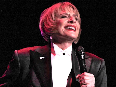 Person in a tuxedo jacket and bow tie holds a microphone and smiles while performing on stage under pink and white lighting.