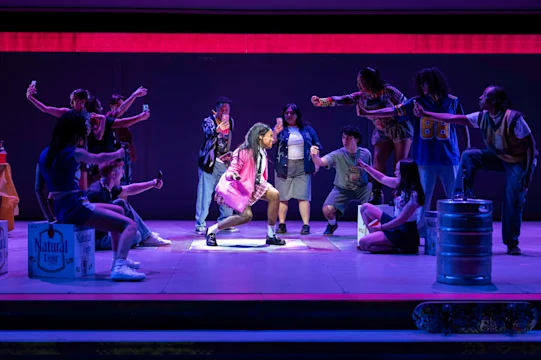 A group of people on stage dramatically surround a person in a pink outfit, some holding drinks and phones, with props like a keg, boxes, and a skateboard visible.