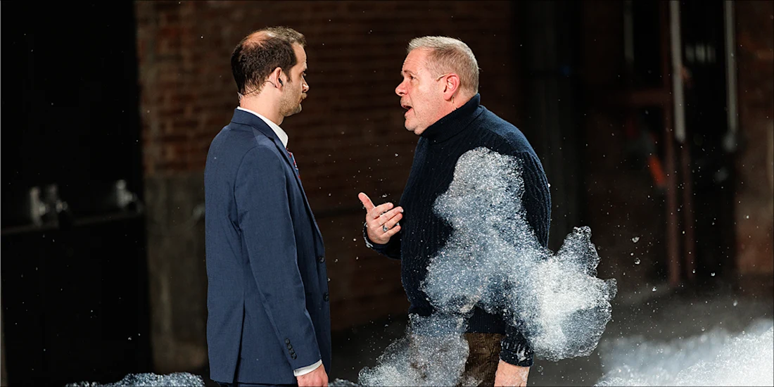 A man in a suit and a man in a sweater talk to each other while surrounded by soap bubbles.