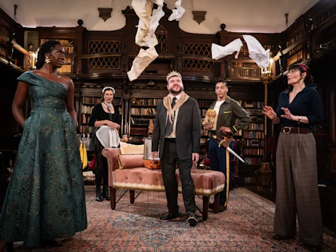 Five actors in costume perform a dramatic scene in a library, with papers flying in the air and various props, including a sword and a crown, on stage.