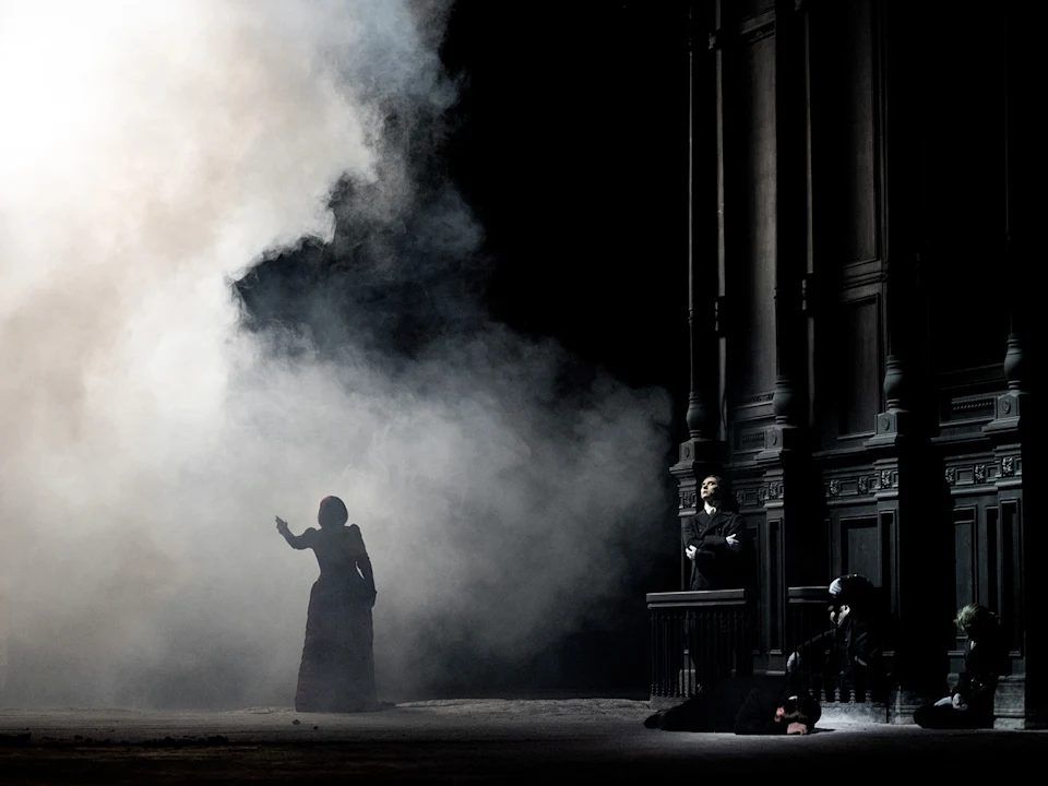 A person in dark clothing stands in fog, gesturing, while others sit by a large building. The scene is dimly lit.