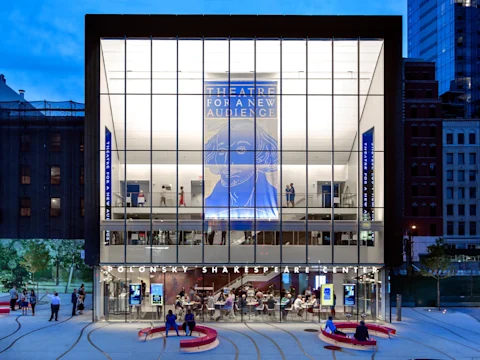 A modern glass-fronted theater building at dusk, with large banners reading “Theatre for a New Audience” and people visible inside.