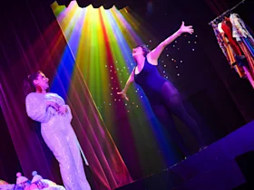 Two performers on stage: one in a sequined outfit looking toward the other, who is in a black leotard with arms outstretched under colorful stage lights. Clothing rack visible on the right.